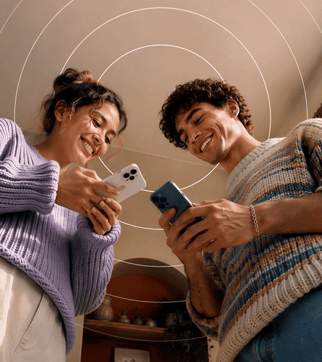 A smiling man and woman in sweaters stand side-by-side, each holding a smartphone beneath a graphic of concentric circles.