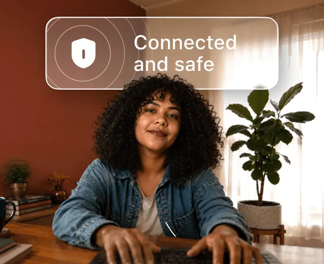A person with curly hair types on a laptop at a desk with a speech bubble above them reading Connected and safe.