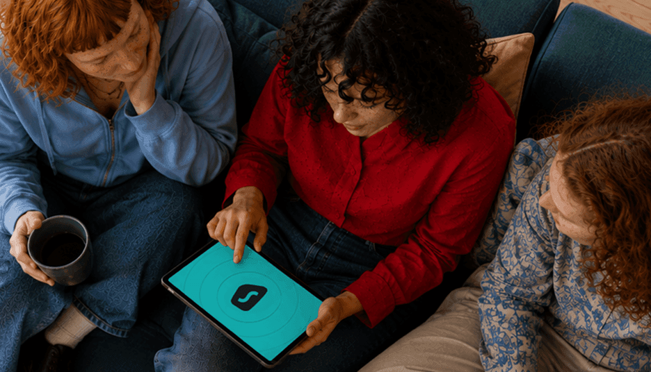 Three people sit on a couch, looking at a tablet screen displaying the Surfshark logo.