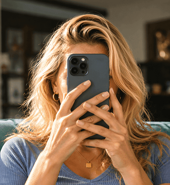 Blonde woman sitting on a couch holding a smartphone in front of her face.